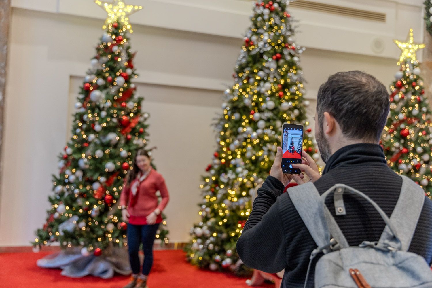 A person takes a photo with a phone of someone posing in front of tall, decorated Christmas trees adorned with lights, ornaments, and glowing star toppers inside a festive indoor display.